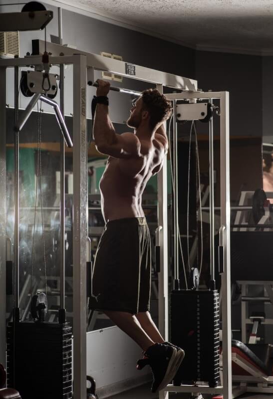 Back view full length muscular shirtless sportsman doing pull ups exercises during workout in contemporary fitness club